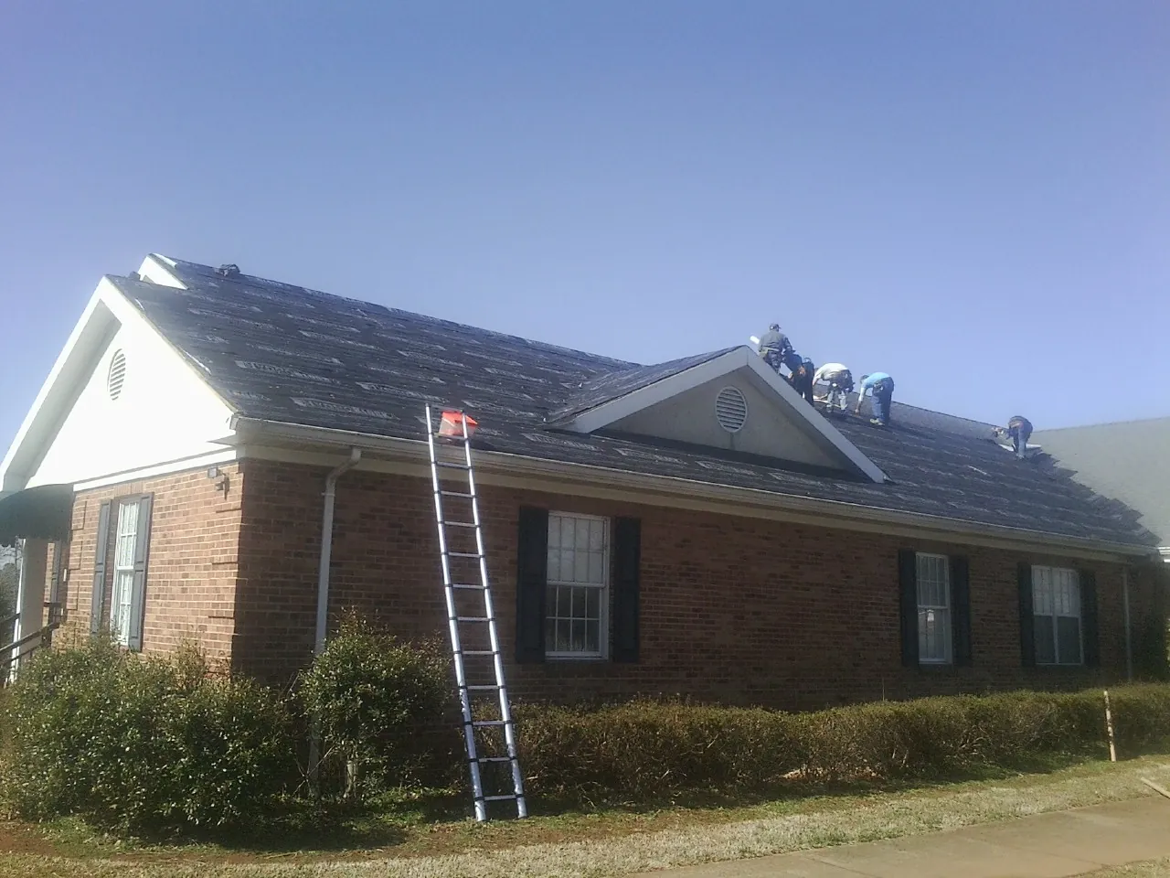 Crew tearing shingles off a brick ranch roof with a ladder set — Metal Roof Repair work by our crew in Lake Grove