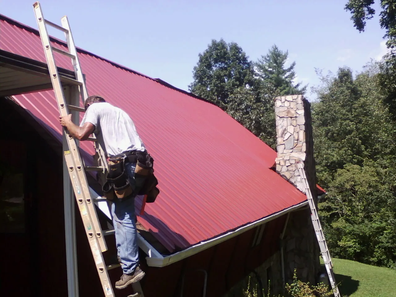 Roofer on a ladder beside a red metal roof and stone chimney — Soffit & Fascia Repair by Roofing Brothers in Lake Grove