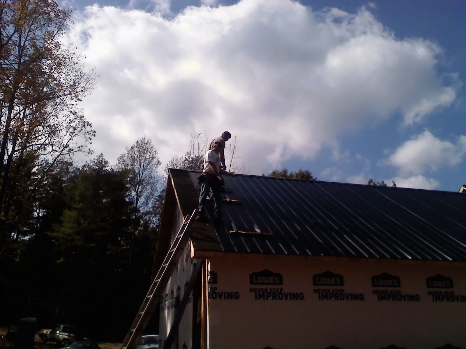 Roofer fastening dark metal panels high against a cloudy sky, from a Roof Inspection job in Lake Grove
